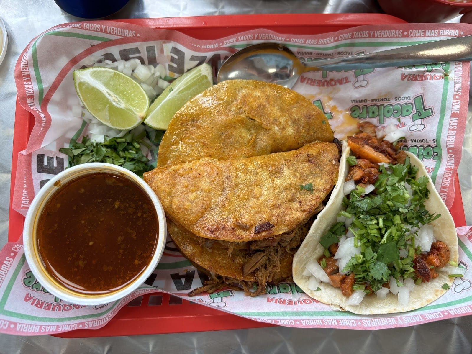 Un plateau contenant des aliments mexicains, dont deux tacos de birria accompagnés de bouillon, d'oignons hachés, de coriandre, de quartiers de citron vert et d'un petit taco. Le plat est garni d'herbes fraîches et une cuillère est placée à côté des aliments.