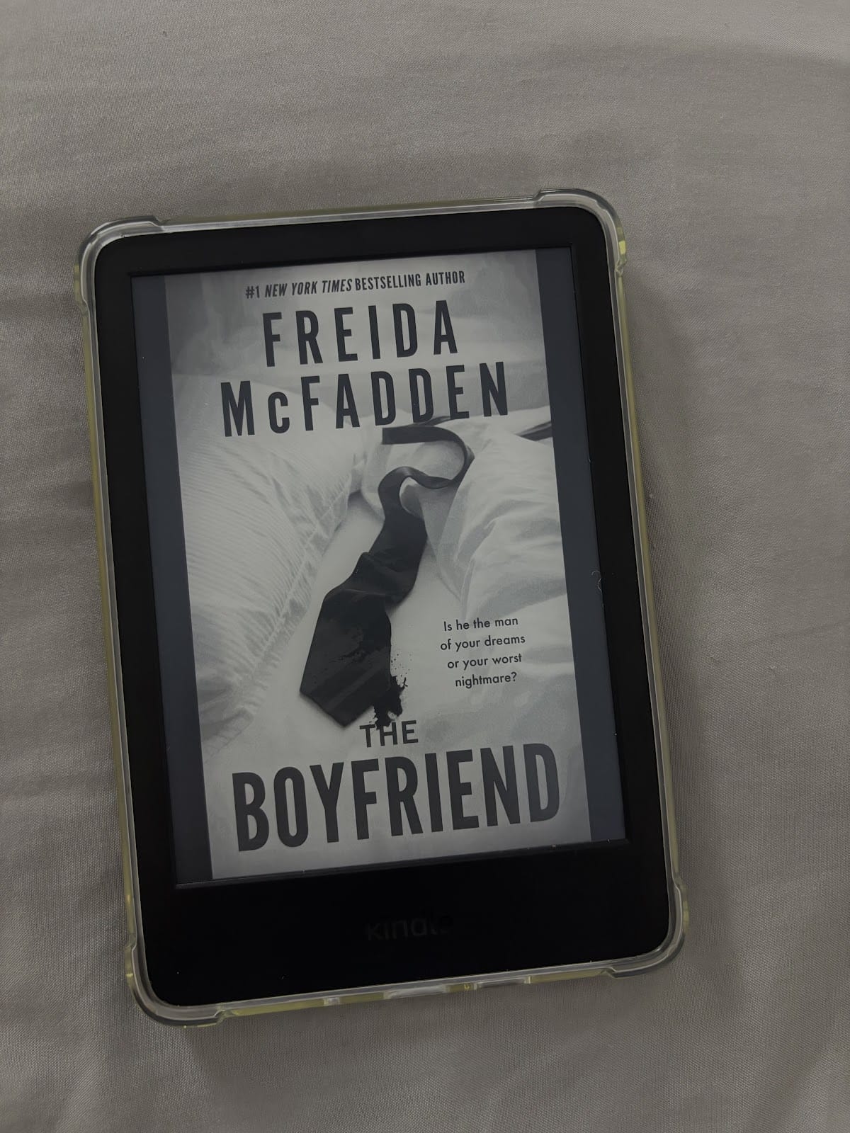 Une liseuse Kindle affichant la couverture du livre "The Boyfriend" de Freida McFadden. La couverture représente une cravate noire sur un couvre-lit blanc, avec le texte suivant : "Est-il l'homme de vos rêves ou votre pire cauchemar ?