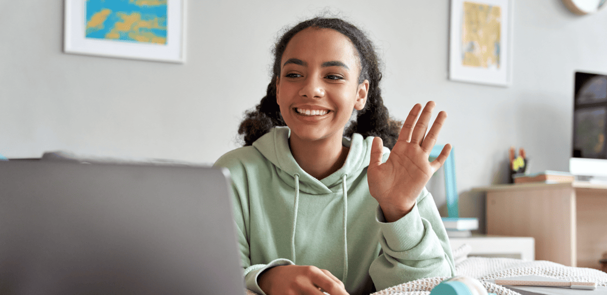 Une jeune femme est assise à un bureau avec son ordinateur portable, souriant et saluant joyeusement la caméra.