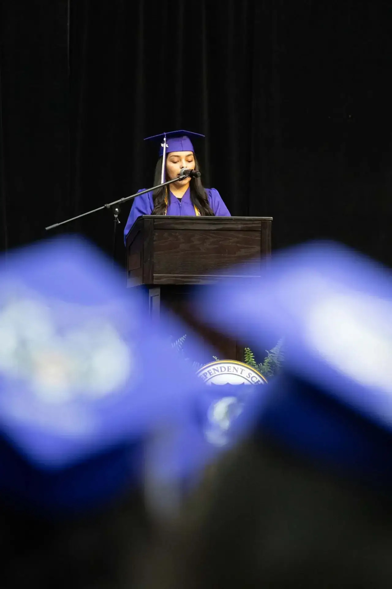 Une femme vêtue d'une robe et d'un bonnet de fin d'études violets se tient sur un podium et parle dans un microphone. Les casquettes bleues des diplômés assis se confondent au premier plan. L'atmosphère est formelle et festive.