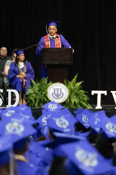 Un diplômé vêtu d'une casquette et d'une toge bleues s'exprime sur un podium lors d'une cérémonie de remise des diplômes. Les diplômés assis et l'emblème de l'école sont visibles, signe de célébration.