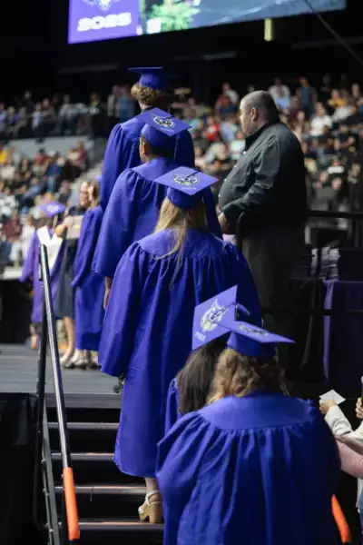 Des diplômés en robe bleue montent sur une scène lors d'une cérémonie, portant des casquettes décorées. Une foule regarde en arrière-plan, créant une atmosphère de fête.