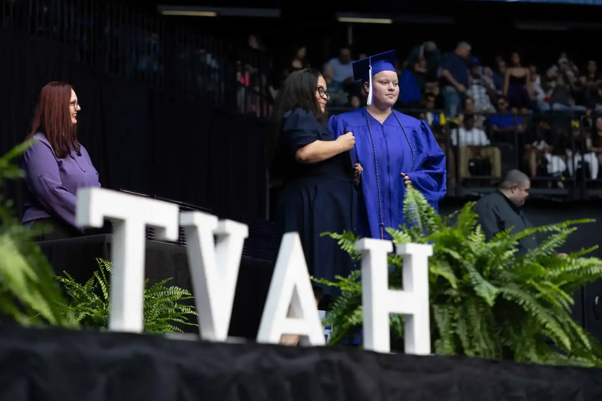 Le diplômé, vêtu d'une casquette et d'une toge bleues, reçoit ses félicitations sur scène, au milieu de fougères luxuriantes et de grandes lettres "TVAH", sous le regard d'un public lors d'une cérémonie officielle.