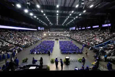 Vue grand angle d'une cérémonie de remise de diplômes dans une arène couverte. Les diplômés, vêtus de casquettes et de toges bleues, sont assis au centre, entourés d'un large public. L'ambiance est formelle et festive.