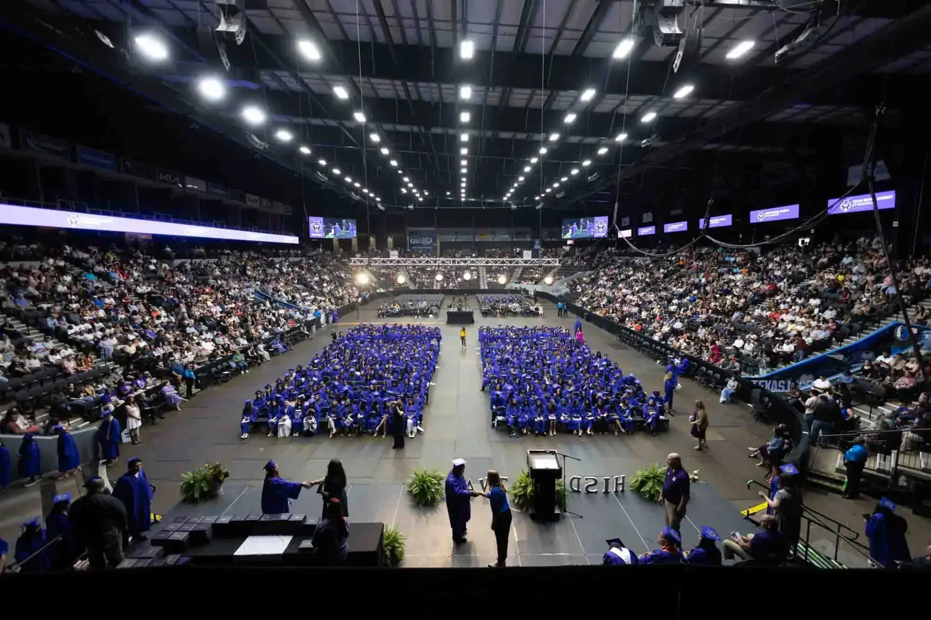 Vue grand angle d'une cérémonie de remise de diplômes dans une arène couverte. Les diplômés, vêtus de casquettes et de toges bleues, sont assis au centre, entourés d'un large public. L'ambiance est formelle et festive.