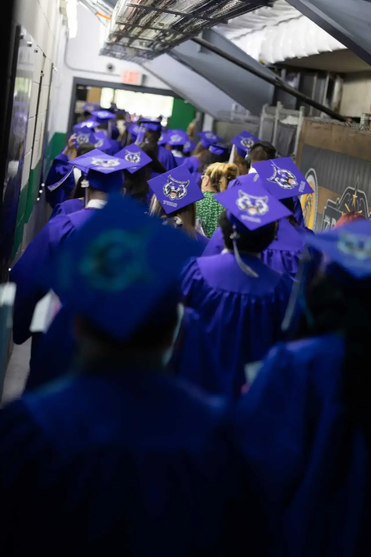 Les diplômés, vêtus de blouses violettes et de mortiers, marchent dans un couloir, l'impatience au cœur. Les casquettes sont ornées de motifs personnalisés, ce qui leur donne un air de fête.