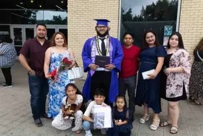 Un diplômé, coiffé d'un bonnet bleu et vêtu d'une toge, tient son diplôme, entouré de sept membres de sa famille qui sourient. Certains tiennent des fleurs et un ours en peluche, fêtant l'événement à l'extérieur.