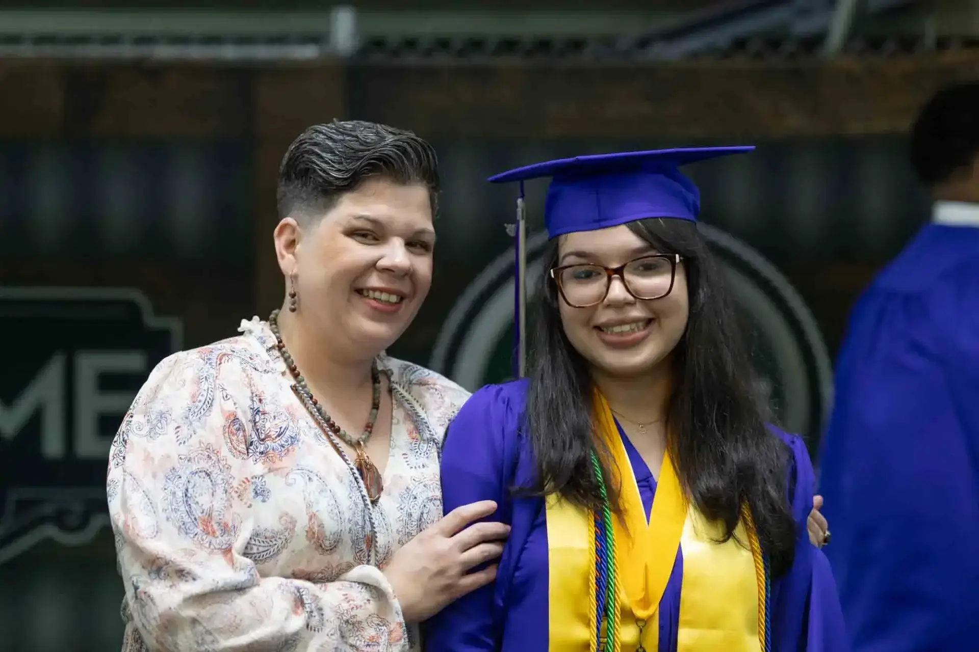 Un diplômé souriant, vêtu d'une toge et d'un bonnet bleus, se tient à côté d'une femme fière aux cheveux courts. La scène transmet la joie et l'accomplissement.