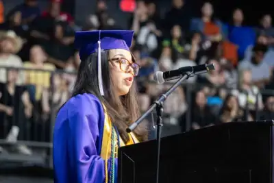 Un diplômé vêtu d'une casquette et d'une toge bleues s'adresse avec assurance à l'auditoire sur un podium. L'arrière-plan montre une foule floue.