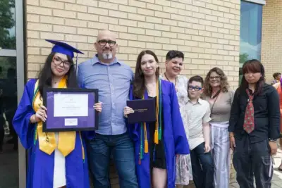 Deux diplômés, vêtus de casquettes et de toges bleues, tiennent leur diplôme en souriant fièrement. Ils sont entourés de membres de leur famille qui les soutiennent, sur fond de mur de briques.