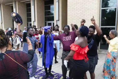 Célébration de la remise des diplômes à l'extérieur d'un bâtiment. Un diplômé vêtu d'un bonnet et d'une toge bleus est joyeusement entouré d'amis et de membres de sa famille qui l'acclament, capturant ainsi un moment plein de vie.